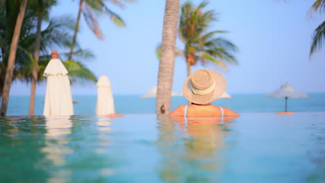 Close-up-of-the-back-and-shoulders-of-a-woman-wearing-a-straw-sun-hat-while-relaxing-on-the-edge-of-a-resort-infinity-edge-pool-looking-out-at-the-beach-and-ocean-beyond