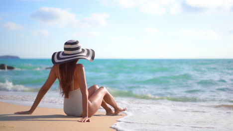 With-her-back-to-the-camera,-a-sexy-young-woman-in-a-bathing-suit-and-floppy-straw-hat-sits-on-a-sandy-beach-enjoying-the-incoming-tide
