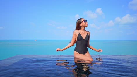 Pretty-woman-in-swimwear-sitting-on-rooftop-infinity-pool-border-and-raising-hands-up-with-a-stunning-view-of-endless-turquoise-seascape-on-horizon-in-Phuket,-Thailand,-static-copy-space