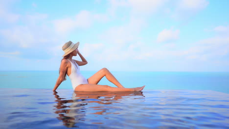 Rooftop-infinity-pool---Woman-in-white-swimwear-and-sunhat-sitting-on-infinity-pool-border,-leaning-on-one-arm-with-a-stunning-view-of-turquoise-sea-water-on-the-horizon-in-Bali,-static