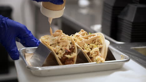Chef-drizzles-sauce-on-top-of-fish-tacos-plated-on-metal-tray-taco-stand-rack,-close-up-slow-motion-4K