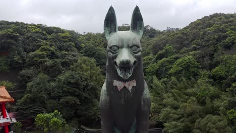 Cinematic-aerial-backwards-shot-showing-gigantic-dog-statue-beside-Asian-temple-in-taiwan-surrounded-by-green-forest-trees-in-the-mountains