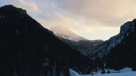 Rising-aerial-drone-shot-of-a-winter-landscape-of-Mount-Timpanogos-in-the-background-surrounded-by-a-pine-tree-forest-during-sunset-from-the-frozen-Tibble-Fork-Reservoir-in-American-Fork-Canyon,-Utah