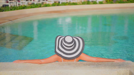 Back-of-a-woman-in-Floppy-striped-hat-leaning-on-concrete-pool-border-inside-swimming-pool,-no-face-template-copy-space