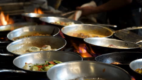Cinematic-view-of-restaurant-stove-covered-with-stainless-skillets-simmering-various-menu-items,-Chef-sautes-in-background,-burner-flames-flare-up-under-pans-of-bubbling-food,-slow-motion-slider-HD