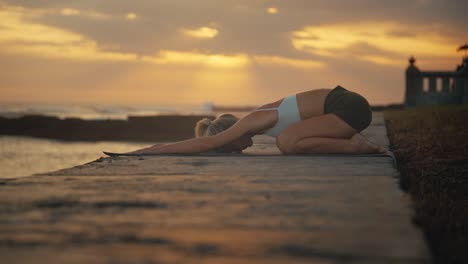 Woman-doing-yoga-child-pose-on-shore-of-Bali-with-sunset-in-background