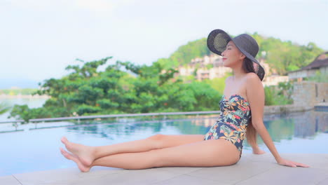Lonely-asian-woman-with-long-legs-sitting-by-the-pool-in-swimsuit-and-floppy-hat-and-enjoying-in-tropical-scenery,-full-frame-slow-motion