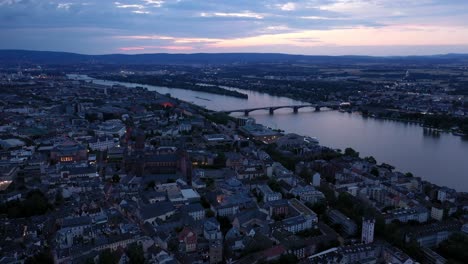 Hohe-Drohne-Luftaufnahme-Von-Mainz-Zur-Magischen-Stunde-Nacht,-Die-Um-Das-Stadtzentrum-Kreist,-Mit-Dem-Dom-Und-Dem-Dunklen-Rheinwasser-Im-Hintergrund,-Das-Einen-Farbenfrohen-Himmel-Zeigt