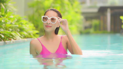 Portrait-of-Wet-Sexy-Asian-Woman-in-White-Sunglasses-and-Bikini-Walking-Inside-Swimming-Pool-and-Looking-at-Camera-Lens,-static-slow-motion