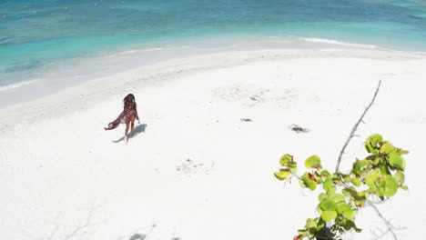 Aerial---Tourist-woman-in-sarong-on-tropical-beach-walks-towards-turquoise-Caribbean-waters