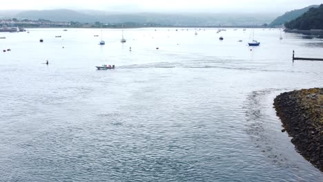 Motorboat-aerial-view-leaving-Welsh-seaside-town-harbour-cruising-along-River-Conwy