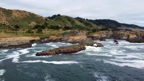 Oregon---Tracking-Back-on-a-coastal-mossy-rock-as-the-ocean-waves-crash-against-oceanside-rock-and-seagull-birds-fly-by-PNW-forests-and-epic-homestead-background,-turquoise-blue-water,-marine-lagoons