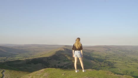 Stabilised-shot-of-young-blonde-woman-standing-on-top-of-Mam-Tor,-Castleton,-Peak-District,-England-admiring-the-view-of-green-rolling-hills-and-blue-skies