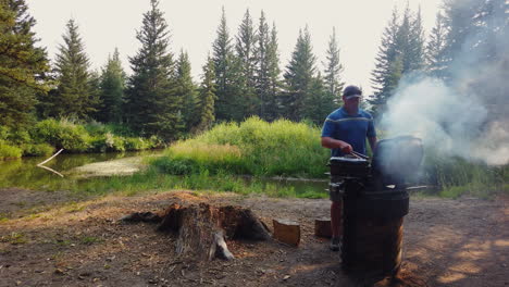Man-Grilling-Steak-On-Riverbank-During-Camping-Trip-In-The-Forest