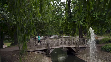 Handheld-panning-shot-of-young-woman-in-green-raincoat-walking-across-wooden-bridge-in-front-of-fountain-in-Weston-Park,-Sheffield,-England