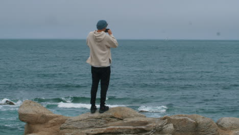 Casual-looking-Young-Man-with-blue-hat-taking-photo-with-smartphone-in-front-of-ocean