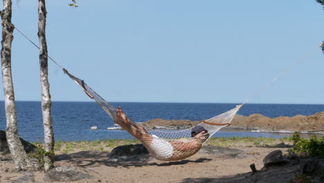Person-sleeping-on-hammock-and-swinging-in-light-breeze-on-ocean-coastline
