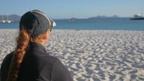Rückansicht-Einer-Brünetten-Frau-Mit-Mütze,-Die-Auf-Dem-Sand-Sitzt-Und-Meerblick-Am-Strand-Von-Whitehaven-In-Queensland-Genießt