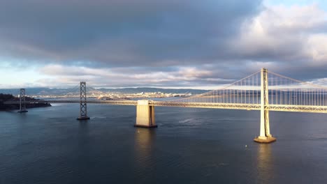 Historische-San-Francisco-Oakland-Bay-Bridge---Autos-Fahren-Entlang-Der-Autobahn,-Stürmische-Kumuluswolken,-Ruhiges-Buchtwasser,-Sonnenuntergang,-Der-Von-Wellen-Reflektiert-Wird,-Filmische-Landschaft,-Die-Zum-Wasser-Hinunterfährt-4k-30fps