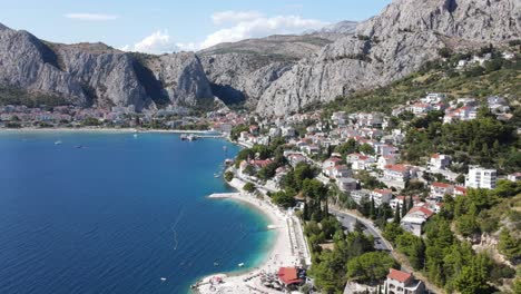 An-aerial-view-over-lush-green-pines-and-steel-grey-mountains-in-the-background,-next-to-white-pebbled-beach-and-deep-blue-Adriatic,-Omis,-Croatia