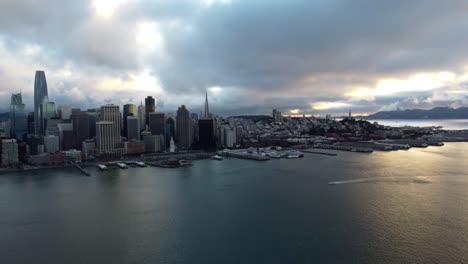 4K-30fps-Aerial-Drone-Footage-of-San-Francisco-Skyline---Small-boat-cuts-through-SF-Bay-water-as-sunset-reflects-off-calm-waves,-beautiful-city-skyline-architecture,-moody-Pacific-Northwest-PNW-sky