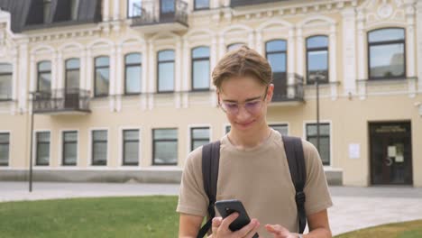 Close-up-shot-of-happy-teenage-boy-walking-from-school-after-lessons-and-smiling-while-texting-with-his-phone