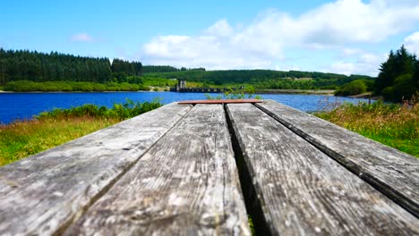 Low-angle-wooden-plank-picnic-table-overlooking-blue-sky-scenic-reservoir-lake-landscape-dolly-right
