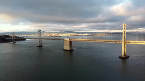 Tracking-In-Aerial-Drone-Footage-of-San-Francisco-Oakland-Bay-Bridge-over-the-SF-Bay---Cars-Road-Trip-Interstate-80,-Sailboats,-Overcast-Cumulus-Clouds,-Calm-Water,-Sun-Reflecting-off-Waves-4K-30fps