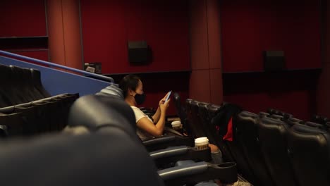 Solo-Girl-In-Facemask-Using-Mobile-Phone-While-Sitting-Alone-In-An-Empty-Movie-Cinema