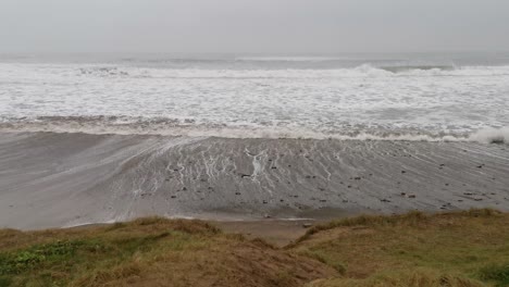 Stormy-day,-high-winds-and-tides,-waves-approaching-a-beach-and-a-patch-go-grass-on-a-sandy-dunes