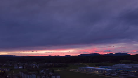 Dark-blue-and-purple-sky,-aerial-hyper-lapse,-timelapse-of-sunset-above-small-town-in-Europe-with-streaks-of-sun-beams-behind-mountains