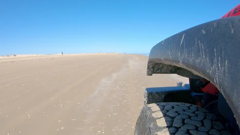 Left-front-tire-of-vehicle-and-sand-dunes-while-driving-on-a-beach-on-a-sunny-day-on-South-Padre-Island-Texas--Point-of-view,-POV