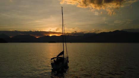 Beautiful-aerial-drone-orbit-around-sail-boat-in-calm-ocean-with-amazing-orange-sunset-with-clouds-and-mountains