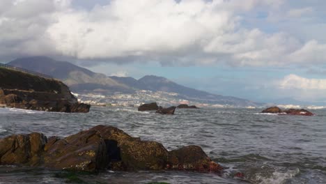 Rocks,-ocean-waves-and-clouds-with-town-on-the-background