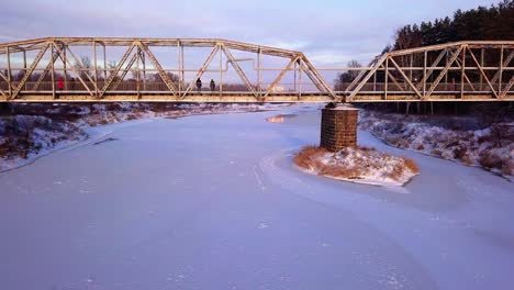 Winter-drone-shot-flying-towards-and-under-an-old-railway-bridge-over-a-ice-covered-river-in-Valmiera,-Latvia