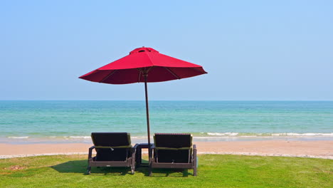 Zwei-Liegestühle-Unter-Sonnenschirm-Auf-Grünem-Gras-Teil-Der-Strandlounge-Am-Meer-Tagsüber
