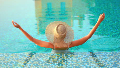 Back-of-the-woman-in-sun-hat-raising-hands-up-while-standing-in-swimming-pool-in-Miami,-elevated-back-view-slow-motion