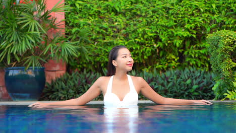 An-attractive-Asian-woman-relaxing-at-a-Hotel-spa,-leaning-on-the-edge-of-a-swimming-pool-on-a-greenery-background,-slow-motion