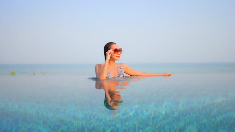 Young-Asian-woman-with-sunglasses-relaxing-in-infinity-pool-and-turquoise-sea-in-background