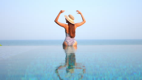 Back-view-of-woman-sitting-on-pool-edge-raising-arms-and-looking-at-horizon-sea