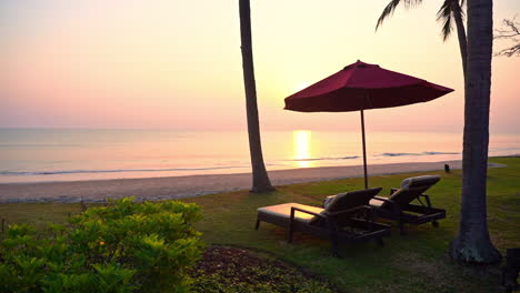 An-empty-pair-of-sun-loungers-under-an-open-beach-umbrella-face-the-sunset-as-ocean-waves-roll-in-along-the-shore