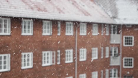 Snowflakes-are-falling-against-blurry,-red-brick-house,-winter-day