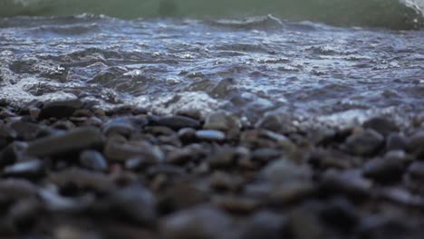 Low-angle-shot-of-small-wave-breaking-on-beach-filled-with-stones,-slow-motion
