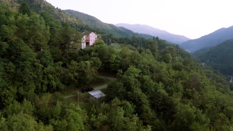 Beautiful-aerial-drone-shot-of-a-large-house-standing-in-the-middle-of-a-forest-on-a-mountain,-black-car-driving-up-a-narrow-road-towards-the-house,-drone-slowly-flying-closer