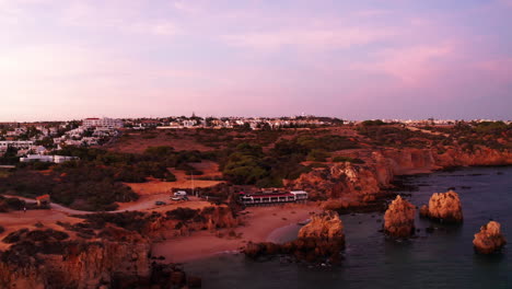 Aerial-shot-of-red-colored-landscape-during-sunset-showing-ocean-water,-beach-and-hotel-resort-on-hilltop