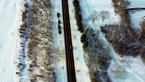 Aerial-Birds-Eye-View-fly-down-winter-road-next-snow-covered-river,-empty-polished-skating-track-and-rink-on-a-sunny-reflective-afternoon-following-grey-suv-down-2-two-lanes-quiet-roadway-park-2-3