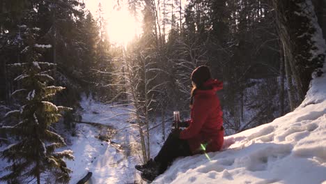 Mädchen-Im-Roten-Wintermantel,-Das-Auf-Einem-Schneebedeckten-Hügel-Sitzt-Und-Ein-Glas-Teeflasche-Hält-Und-Das-Helle-Sonnenlicht-Bewundert