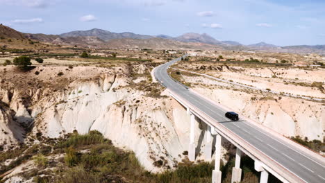 Drone-capturing-a-beautiful-scenery-of-a-black-car-driving-over-a-bridge-in-the-middle-of-a-stunning-landscape-made-out-of-mountains-and-a-mixture-of-grassland-and-savanna,-Murica,-Spain