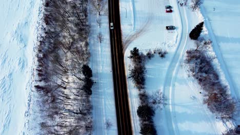winter-aerial-birds-eye-view-2-two-lane-roadway-next-to-snow-covered-park-river-and-empty-quiet-skating-track-and-ice-rink-with-bushes-and-trees-around-it-and-a-few-parked-cars-following-white-van-3-3