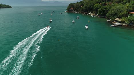 Aerial-drone-shot-of-lots-of-speedboat-passing-through-moored-boats-near-the-coastline-of-a-tropical-forest-with-dense-vegetation-and-few-constructions